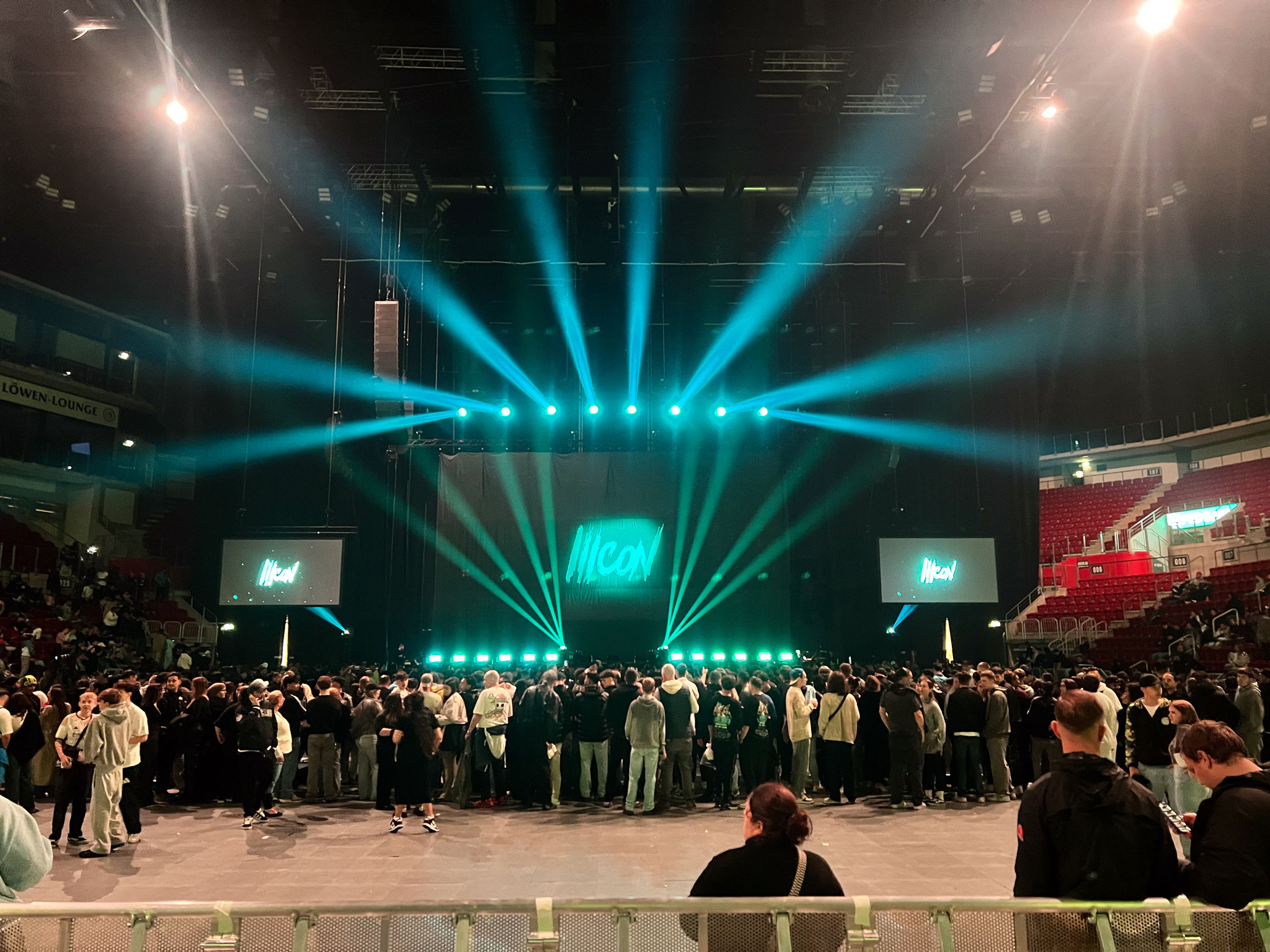 Sehr große Bühne in einer dunklen Halle mit vielen Menschen und türkisfarbenen Lichtstrahlen.