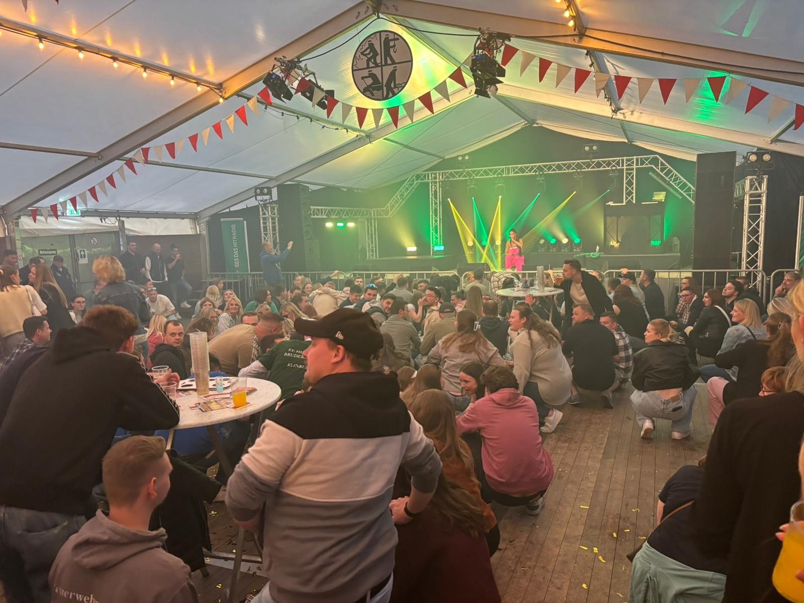 Große Menschenmenge, die vor der Bühne in einem Festzelt in der Hocke sitzt.