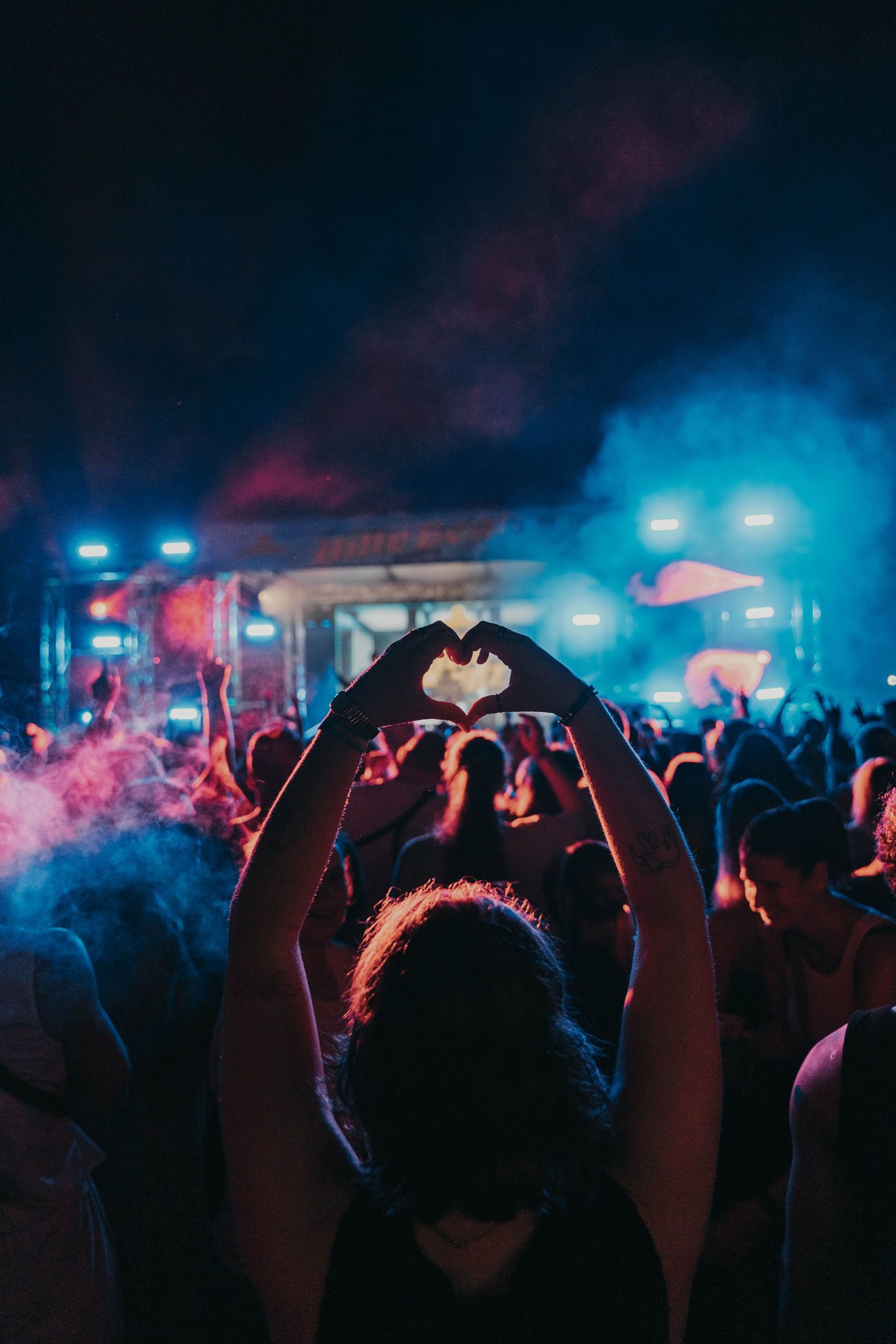 Eine junge Frau formt mit ihren Händen ein Herz in einer tanzenden Menge vor einer bunt beleuchteten Bühne im Freien.