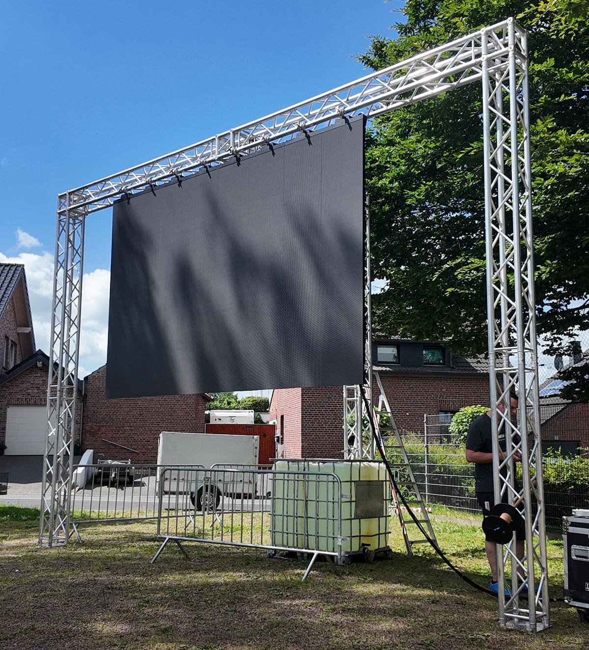 Große Traversenkonstruktion mit einer mittig abhängenden LED-Videowand in einer kleinen Parkanlage.