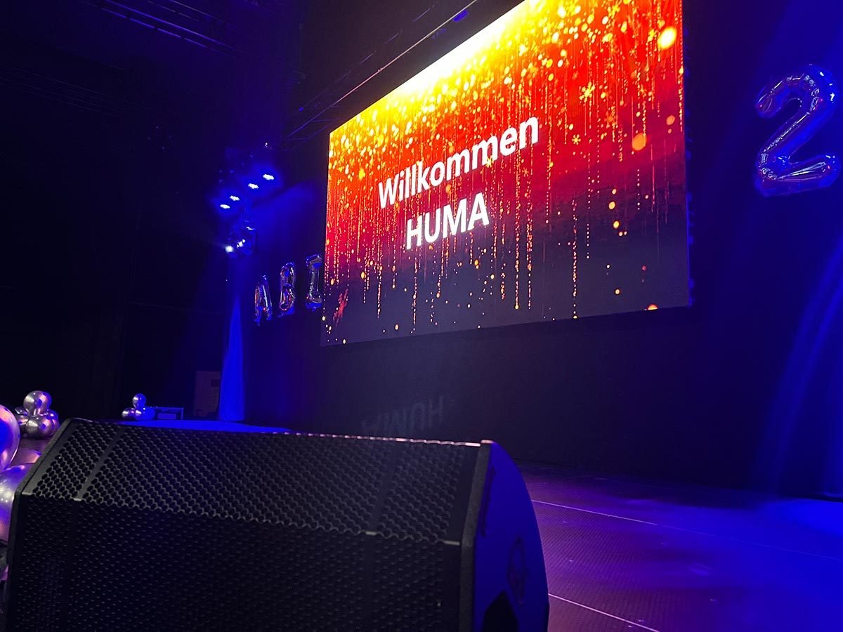 Seitliche Perspektive einer dunklen Halle mit großer, blau beleuchteter Bühne und heller LED-Videowand im Hintergrund.
